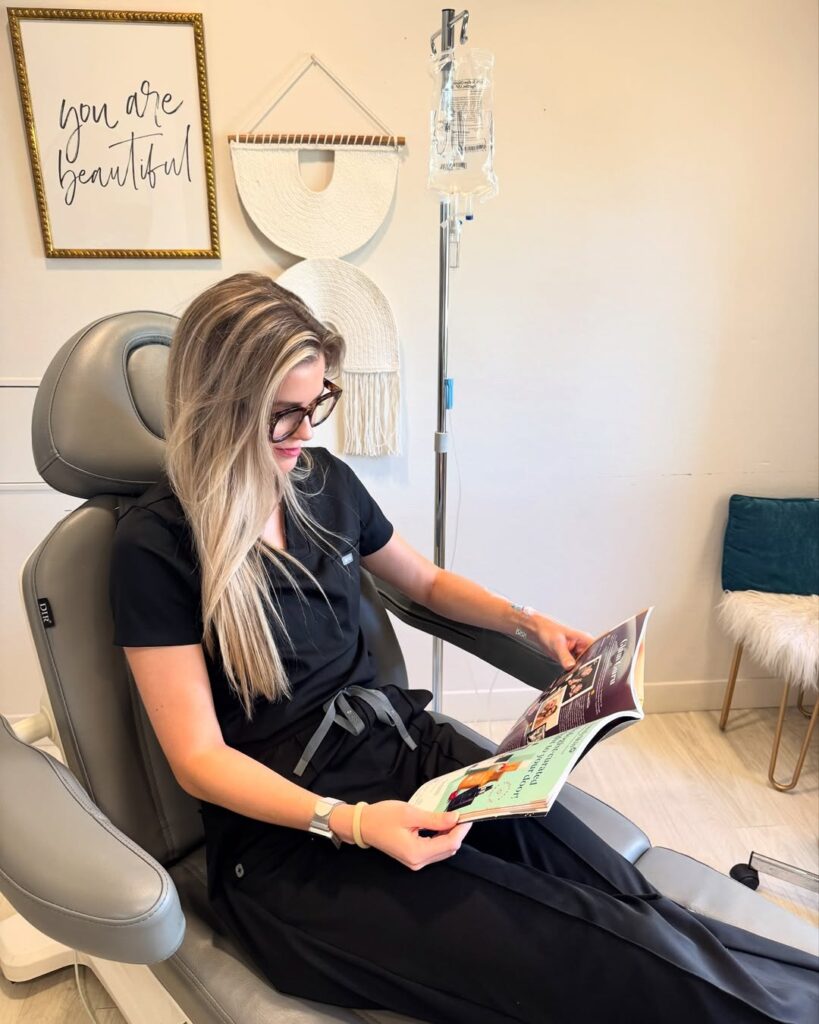 A woman in black scrubs reading a magazine while receiving a vitamin drip via IV therapy at a wellness spa.