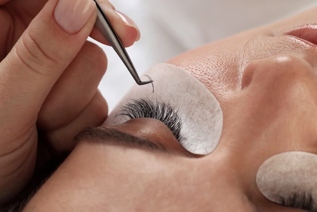A close-up of a beauty technician using tweezers to carefully apply an individual eyelash extension to a client's natural lashes.