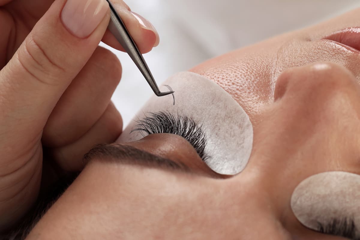A close-up of a beauty technician using tweezers to carefully apply an individual eyelash extension to a client's natural lashes.